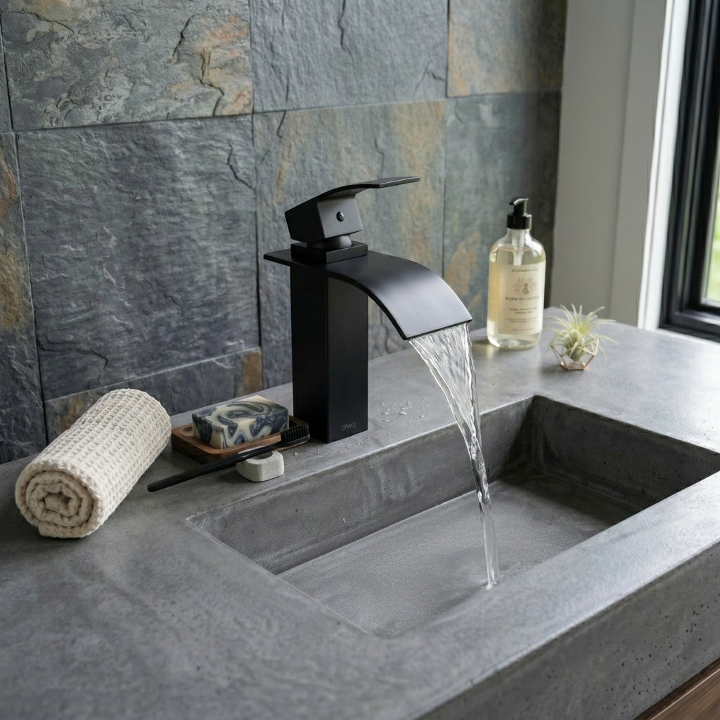 Onyx Elements matte black waterfall bathroom faucet with single handle on a grey concrete vanity with stone wall.